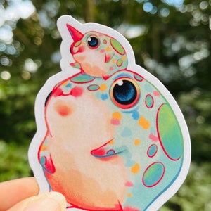 May include: A sticker featuring two cartoon frogs. The larger frog is green, pink, and orange with white spots. The smaller frog is on top of the larger frog and is white with green and pink spots. Both frogs have large, black eyes.