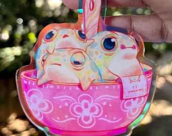 May include: A pink ceramic bowl with a floral design, containing three cartoon frogs. The bowl has a spoon sticking out of it. The frogs are white with colorful spots and have large eyes. The bowl has a small white tag with the text "ES MONKEY" on it.