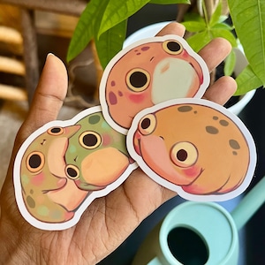 May include: Three frog stickers with different colors and patterns. The stickers are on a hand, and the hand is holding a watering can.