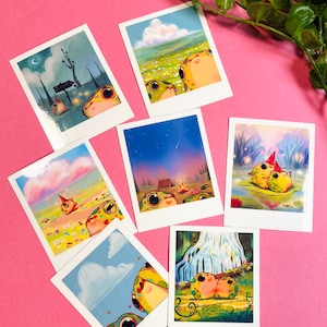 Puede incluir: Conjunto de 7 tarjetas ilustradas de estilo Polaroid con ranas de dibujos animados en diferentes escenarios. Las ranas son verdes con manchas amarillas y tienen ojos grandes. Los escenarios incluyen un bosque, un campo de flores, una fogata, una cascada y un cielo nocturno con una estrella fugaz.