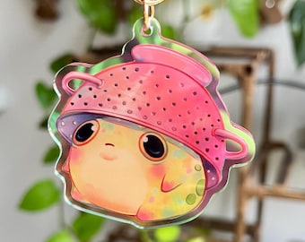 May include: A whimsical keychain featuring a cartoon frog wearing a pink colander. The frog has large, expressive eyes and a cheerful expression. The keychain is attached to a gold-colored ring, perfect for keys or bags.