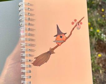 Puede incluir: Un cuaderno de espiral con una cubierta de color melocotón que presenta la ilustración de una pequeña criatura naranja con un sombrero puntiagudo oscuro, montada en una escoba de madera con un hatillo y flores rojas en el mango.