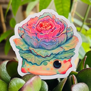 Puede incluir: Una pegatina con una criatura de dibujos animados con una col rizada rosa, azul y verde en la cabeza. La criatura tiene un cuerpo de color naranja claro con un ojo negro y una pequeña boca.