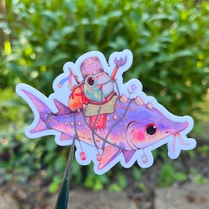 May include: A whimsical sticker featuring a purple fish with pink accents, carrying a frog-like creature wearing a hat that says "BEANS." The frog has a backpack and is holding a spear. The sticker has a white border and is set against a blurred green background.