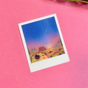 May include: A Polaroid photo of two cartoon frogs sitting in front of a campfire with a tent behind them. The sky is blue with a shooting star and the background is a forest.