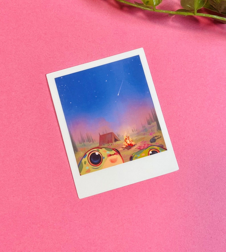 May include: A Polaroid photo of two cartoon frogs sitting in front of a campfire with a tent behind them. The sky is blue with a shooting star and the background is a forest.