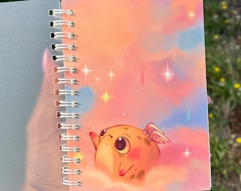 Puede incluir: Un cuaderno de espiral con una ilustración en la portada de una criatura redonda de color naranja con alas pequeñas, flotando en un cielo nublado rosa y azul lleno de estrellas brillantes blancas y amarillas. La cubierta es de plástico translúcido.