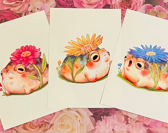 May include: Three watercolor paintings of frogs wearing flowers on their heads. The frogs are all different colors and have different expressions. The flowers are yellow, pink, and blue.