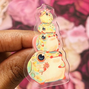 May include: A stack of four colorful frog stickers with a flower on the top frog. The frogs are all different colors and have black eyes. The stickers are on a clear background.