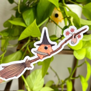 Puede incluir: Una pegatina de una rana de dibujos animados con un sombrero de bruja y montando una escoba. La rana es naranja con una barba marrón y sostiene un palo con flores rosas.