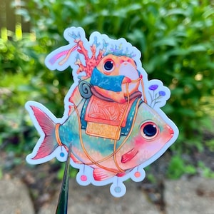 May include: A whimsical sticker featuring a colorful fish with a coral headdress and a backpack. The fish has a teal body, pink fins, and large, expressive eyes. The sticker has a white border and is set against a blurred green background.