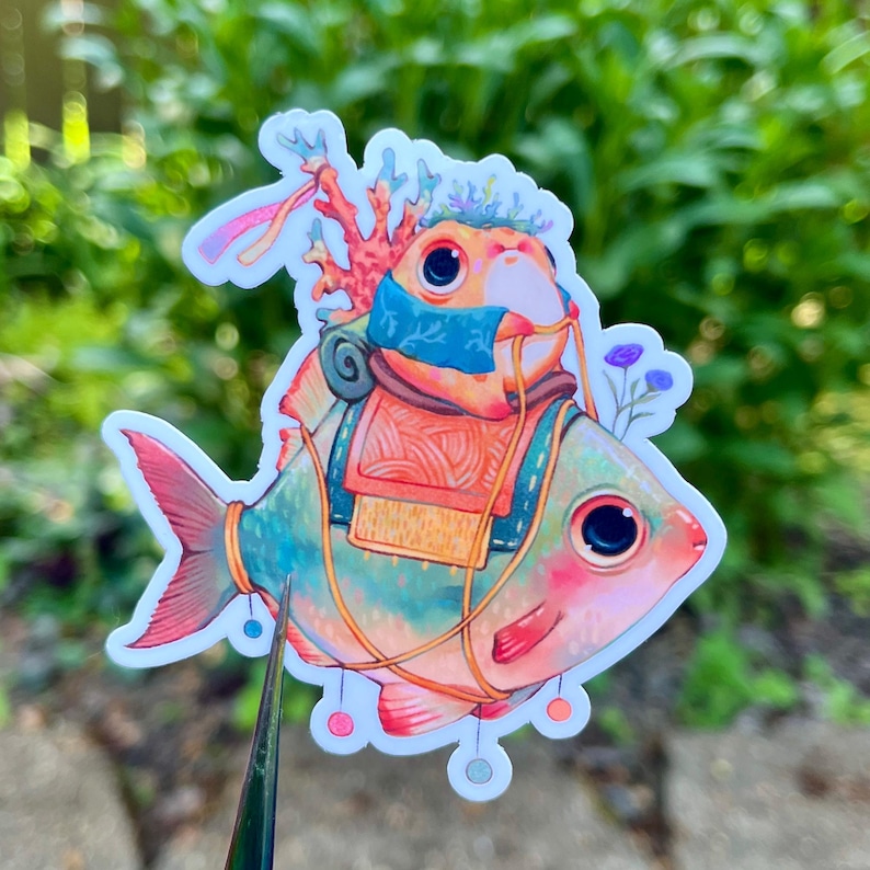 May include: A whimsical sticker featuring a colorful fish with a coral headdress and a backpack. The fish has a teal body, pink fins, and large, expressive eyes. The sticker has a white border and is set against a blurred green background.
