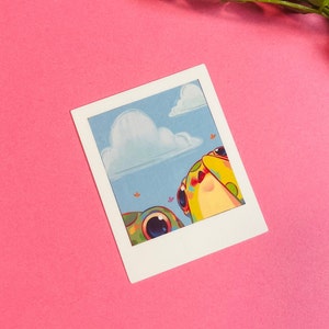 May include: A Polaroid-style photo print featuring two cartoon frogs looking up at a fluffy white cloud in a blue sky. The frogs are green, yellow, and orange with black eyes.
