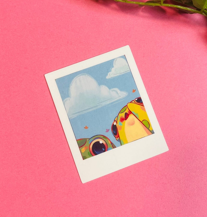 May include: A Polaroid-style photo print featuring two cartoon frogs looking up at a fluffy white cloud in a blue sky. The frogs are green, yellow, and orange with black eyes.