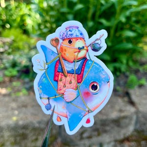 May include: A whimsical sticker featuring a cartoon fish with a blue body and a round eye. The fish is adorned with a small orange character wearing a blue hat and a red and white outfit. A spoon and a small teapot are also included in the design.
