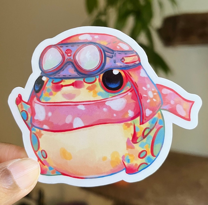 May include: A colourful frog wearing a pink scarf and goggles. The frog has a white background with a blue outline.