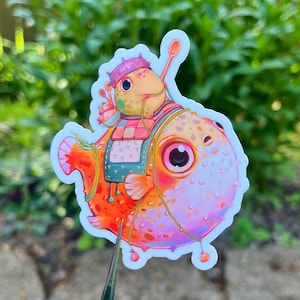 May include: A colorful sticker featuring a cartoon pufferfish character. The fish is orange, pink, and yellow, wearing a chef's hat that says "Soup Gang". It has a small apron and is holding a spoon. The sticker has a white border.
