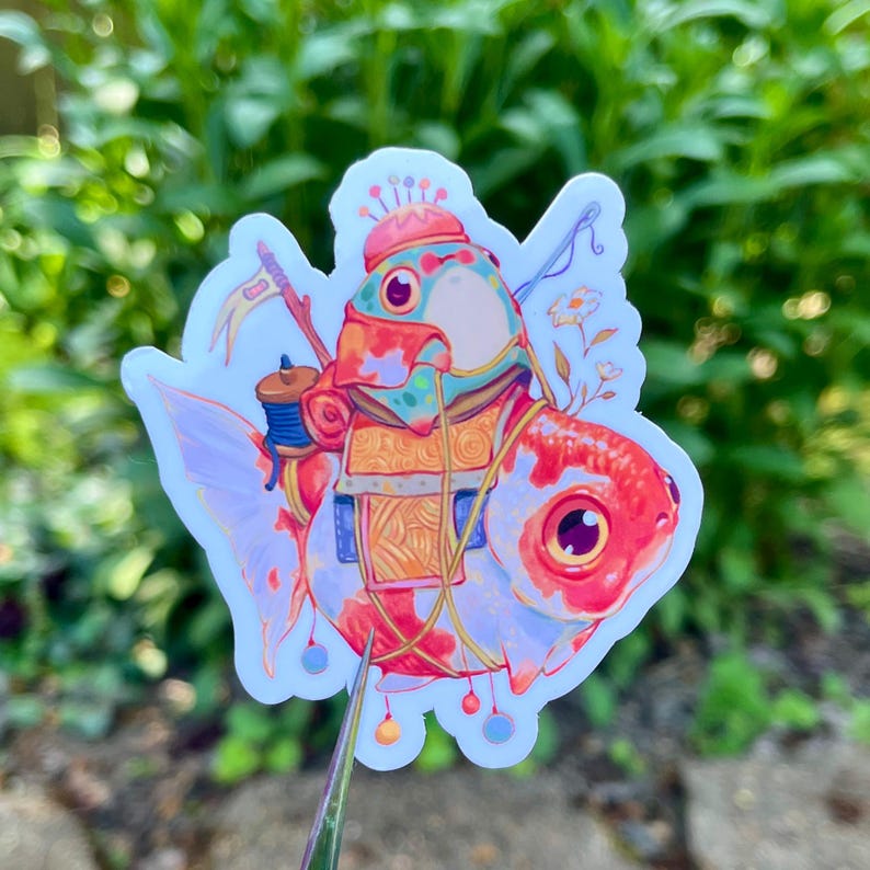 May include: A colorful sticker featuring a whimsical goldfish character. The fish is adorned with a hat, backpack, and other decorative elements. The sticker has a white border and is set against a blurred green background.