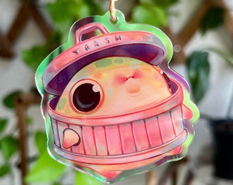 May include: A clear acrylic charm featuring a cartoon character inside a pink trash can. The trash can has the word "TRASH" on the lid. The character has large eyes and a smiling expression. The charm is attached to a gold-colored chain.