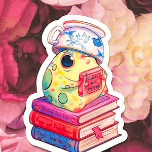 Op de afbeelding: Een kleurrijke kikker met een theekopje op zijn kop zit op een stapel van drie boeken. De boeken zijn getiteld "Teas of the Fen", "Frogs and Fine Brews" en "Toad's Teatime Tips". De kikker houdt een boek vast met de titel "How to Be A Tea Shob".