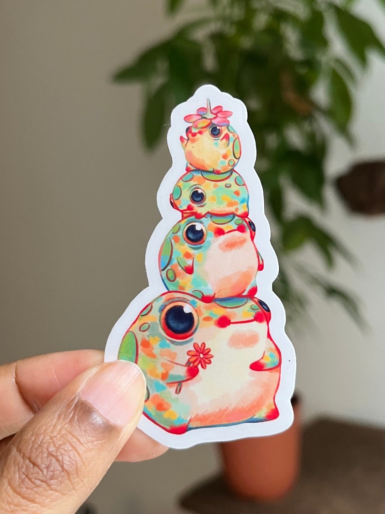 May include: A stack of four colorful frog stickers with a flower on top. The frogs are all different colors and have black eyes. The sticker is on a white background.
