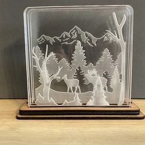 May include: A clear acrylic block with a laser-cut design of a mountain scene with deer and trees. The block is on a wooden base.