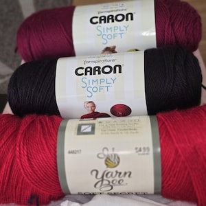 May include: Three skeins of yarn in shades of red, black, and burgundy. The top two skeins are labeled "Caron Simply Soft." The bottom skein is labeled "Yarn Bee Soft Secret."