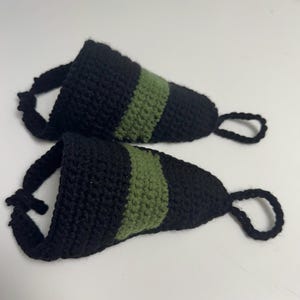 Puede incluir: Dos artículos de ganchillo negros con una franja verde. Cada artículo tiene un lazo en un extremo y una correa en el otro. Los artículos están hechos a mano y parecen ser un tipo de accesorio.