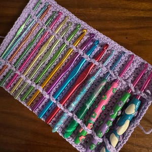 May include: A collection of crochet hooks in various colors, neatly organized in a lavender crochet case. The hooks are made of metal and have different handle designs, including floral and abstract patterns. The case is made of a soft, woven material.
