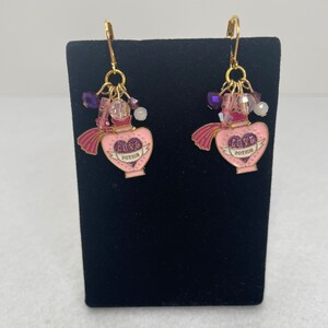May include: A pair of gold earrings with pink heart-shaped charms that say "Love Potion". The charms are decorated with purple glitter and have a pink tassel. The earrings are adorned with clear and purple beads.