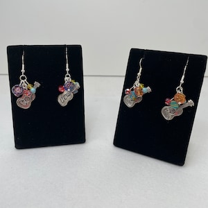 May include: A pair of silver dangle earrings with a guitar charm and colourful beads. The earrings are displayed on black velvet earring stands.