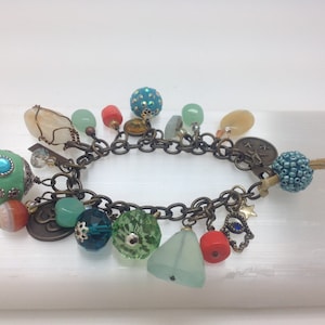 May include: A charm bracelet with a variety of colorful beads, including green, blue, and red. The bracelet is made of metal and has a chain link design. The charms include a star, a hand with an eye, and a small bell.