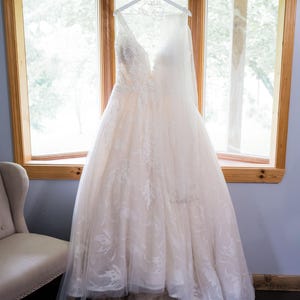 May include: A white wedding dress with lace detailing hangs on a white hanger with the text "06.23.23 Mrs. Amigh" on it. The dress is in front of a window with a view of trees.