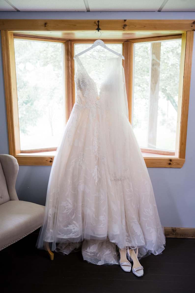 May include: A white wedding dress with lace detailing hangs on a white hanger with the text "06.23.23 Mrs. Amigh" on it. The dress is in front of a window with a view of trees.
