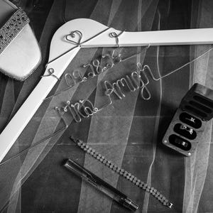 May include: A white hanger with a wire inscription that reads "09.28.24 Mrs. Amy" and two hearts. A pair of sparkly silver shoes, a small wooden box with rings inside, and a silver chain bracelet are also visible.