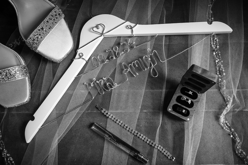 May include: A white hanger with a wire inscription that reads "09.28.24 Mrs. Amy" and two hearts. A pair of sparkly silver shoes, a small wooden box with rings inside, and a silver chain bracelet are also visible.