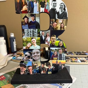 May include: A large wooden letter "B" decorated with multiple photographs. The letter is black and sits on a black base. The photos are of various people, likely family or friends, and are in color. The background is a neutral color.