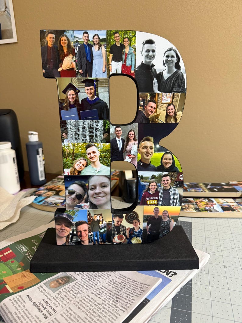 May include: A large wooden letter "B" decorated with multiple photographs. The letter is black and sits on a black base. The photos are of various people, likely family or friends, and are in color. The background is a neutral color.