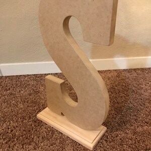 May include: A large, unfinished wooden letter "S" stands upright on a small, rectangular wooden base. The letter is light brown and has a cutout in the center. The base is also light brown and has a decorative edge. The item is likely for home decor.