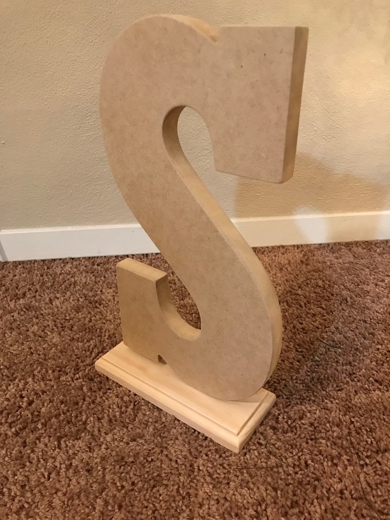 May include: A large, unfinished wooden letter "S" stands upright on a small, rectangular wooden base. The letter is light brown and has a cutout in the center. The base is also light brown and has a decorative edge. The item is likely for home decor.