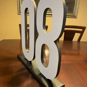 May include: A large, freestanding number "08" in white with black edging, mounted on a black rectangular base. The numbers are displayed on a dark wooden table, suitable for event decor or home accents.