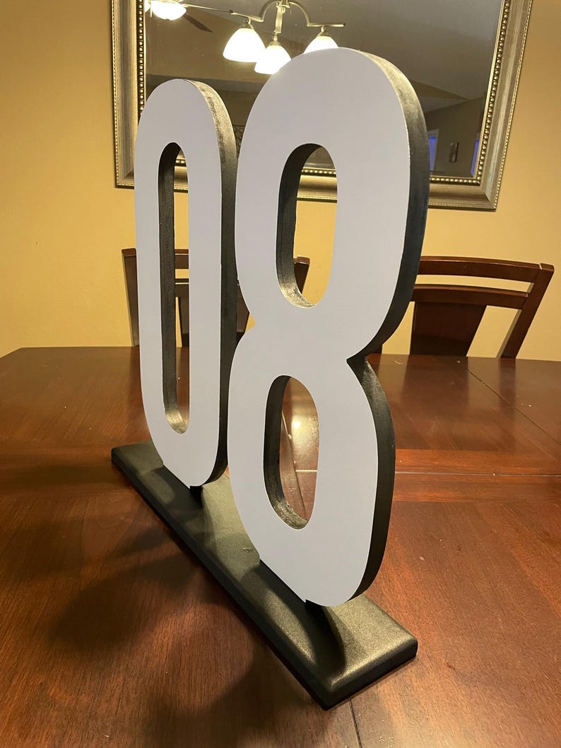 May include: A large, freestanding number "08" in white with black edging, mounted on a black rectangular base. The numbers are displayed on a dark wooden table, suitable for event decor or home accents.