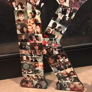 May include: A large wooden letter "K" decorated with numerous photographs. The photos are of various sizes and feature people of different ages and settings. The letter is black with a dark brown border and sits on a light-colored surface.