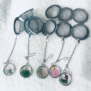May include: Five silver-toned metal tea infusers with mesh spheres and attached chains. Each chain ends in a circular charm containing pressed dried flowers in various colors including white, green, pink, yellow, and purple, encased in clear resin.