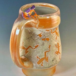 May include: Handmade ceramic mug with an orange handle and rim, featuring orange frog designs on a textured, off-white background. A small, purple and orange frog figurine sits on the rim. A unique, artistic piece for beverages.