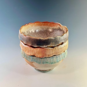 May include: Stacked ceramic bowls with textured, ridged sides and scalloped rims. The bowls are in shades of orange, teal, and gray, with a speckled interior. The handmade pottery has a rustic, artisanal aesthetic.