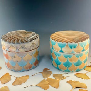May include: Two ceramic lidded jars with a decorative scale pattern in shades of blue, orange, and cream. The lids have a textured, concentric design. The jars are round and sit on a white surface with scattered golden leaves.