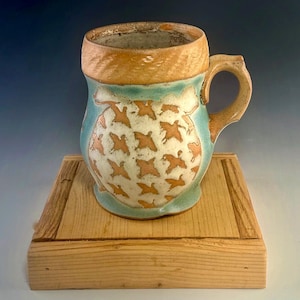 May include: Handmade ceramic mug with a unique design. The mug features a light blue and tan color scheme, with a pattern of orange birds on a white background. The handle and rim have a textured appearance. The mug is displayed on a wooden stand.
