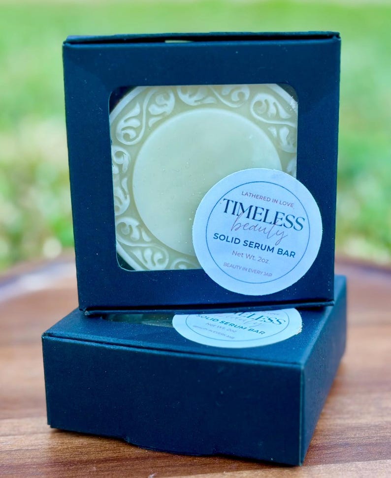 May include: A dark blue box with a clear window displaying a round, light green solid serum bar. The bar is surrounded by a white, ornate design. A sticker on the box reads "TIMELESS beauty SOLID SERUM BAR, Net Wt. 2oz."