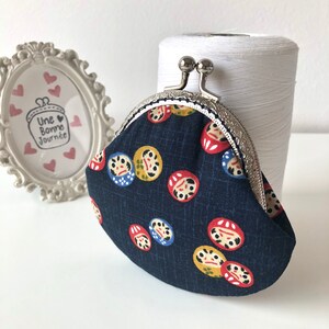 May include: A small, dark blue coin purse with a silver clasp. The purse is decorated with a repeating pattern of red, yellow, and blue Japanese Daruma dolls.